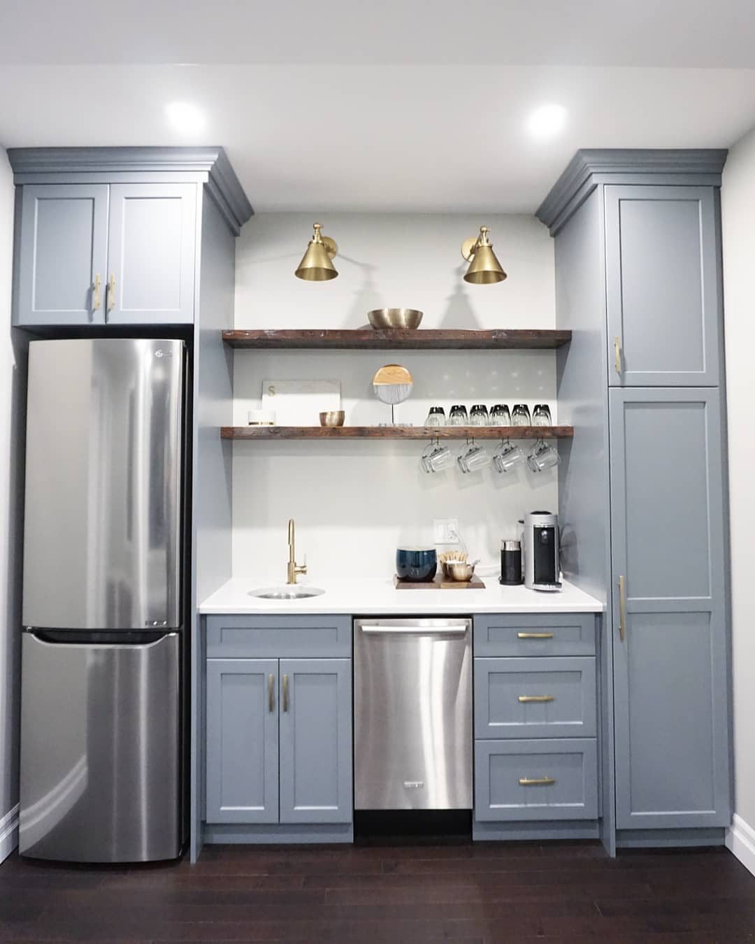 Small blue kitchenette with white countertops, stainless steel appliances, and wooden floating shelves on a white wall.