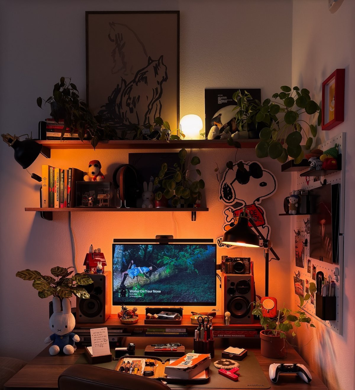 A cozy desk setup with a computer, speakers, many indoor plants, and various small decorative figurines.