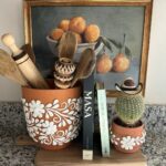 Terracotta pots with white floral designs holding wooden spoons and a small cactus on a wooden board.