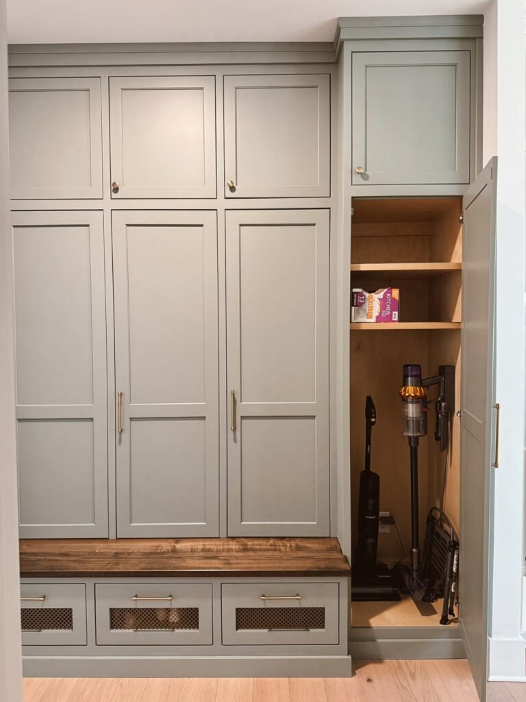 Grey built-in mudroom cabinetry with a wooden bench and an open storage closet containing a vacuum.
