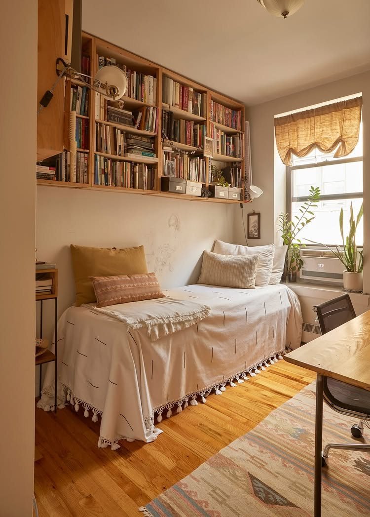 A daybed with white linens sits beneath a large wooden bookshelf in a bright room.